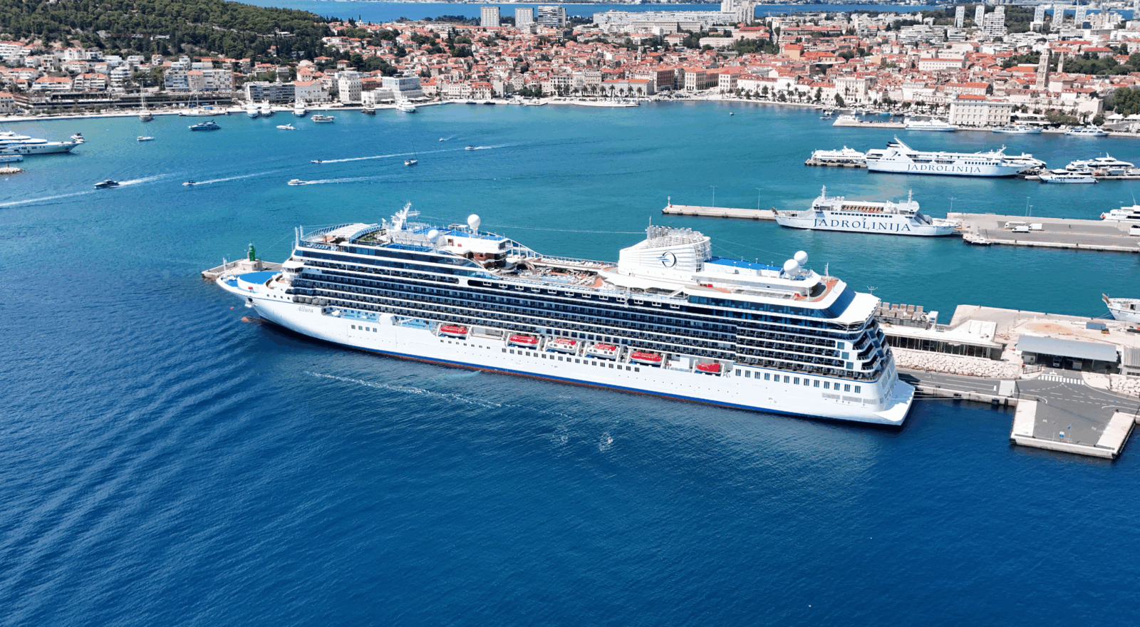 Large white luxury cruise ship docked at the port of Split, Croatia, with turquoise Adriatic waters, Jadrolinija ferries, and the historic old town with red rooftops in the background.