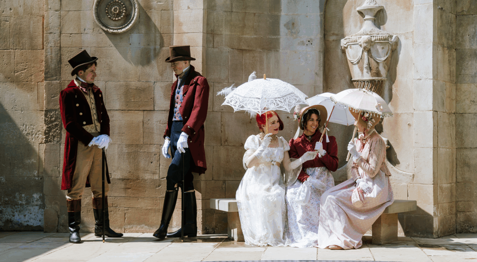 Five people dressed in Regency-era costumes pose outside a stone building, with two men in velvet tailcoats and top hats standing nearby while three women in long gowns hold lace parasols, evoking a Jane Austen festival or historical reenactment.