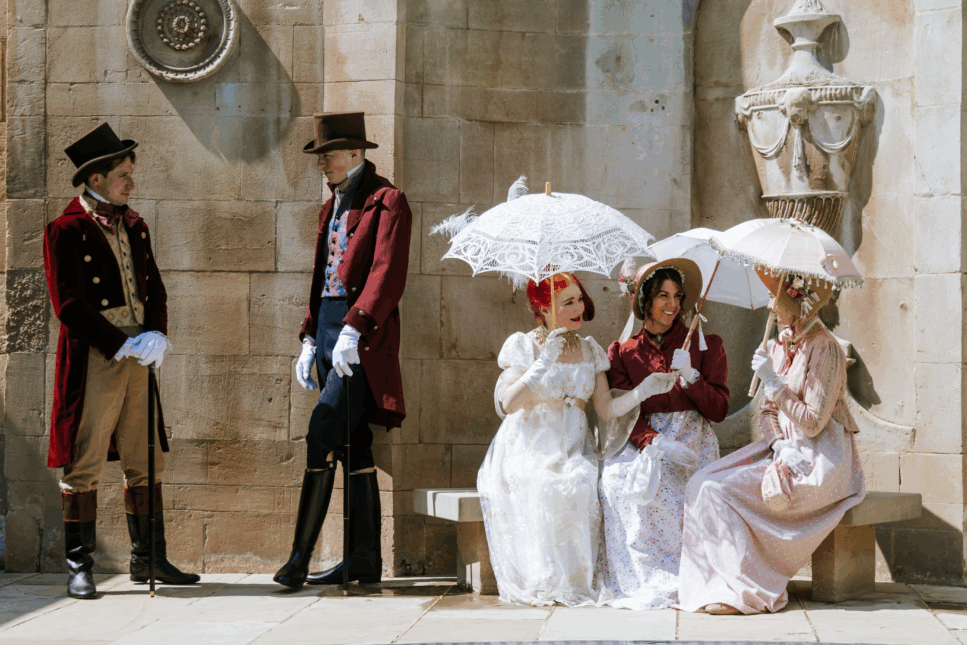 Five people dressed in Regency-era costumes pose outside a stone building, with two men in velvet tailcoats and top hats standing nearby while three women in long gowns hold lace parasols, evoking a Jane Austen festival or historical reenactment.