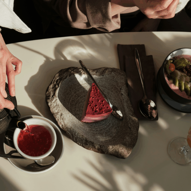 Hands pour herbal tea into a white cup beside a rustic stone plate with raspberry cheesecake, a smoothie bowl topped with fresh fruit and granola, and a glass of citrus-infused drink on a café table with natural sunlight and shadows.