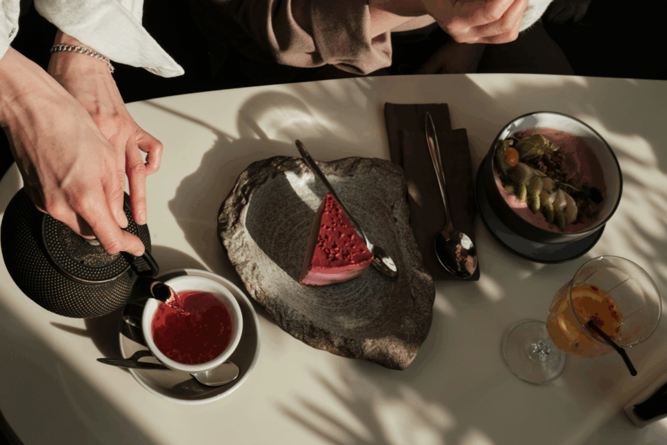 Hands pour herbal tea into a white cup beside a rustic stone plate with raspberry cheesecake, a smoothie bowl topped with fresh fruit and granola, and a glass of citrus-infused drink on a café table with natural sunlight and shadows.