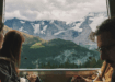 Three travelers sit inside a train with large windows, gazing at a dramatic alpine landscape of snow-capped peaks, glaciers, and green valleys outside.