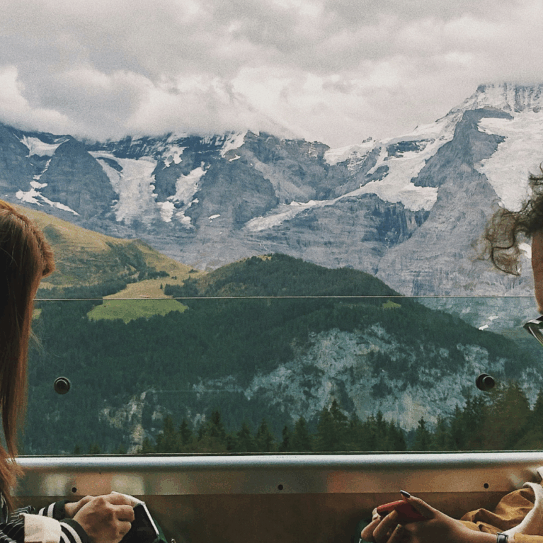 Three travelers sit inside a train with large windows, gazing at a dramatic alpine landscape of snow-capped peaks, glaciers, and green valleys outside.