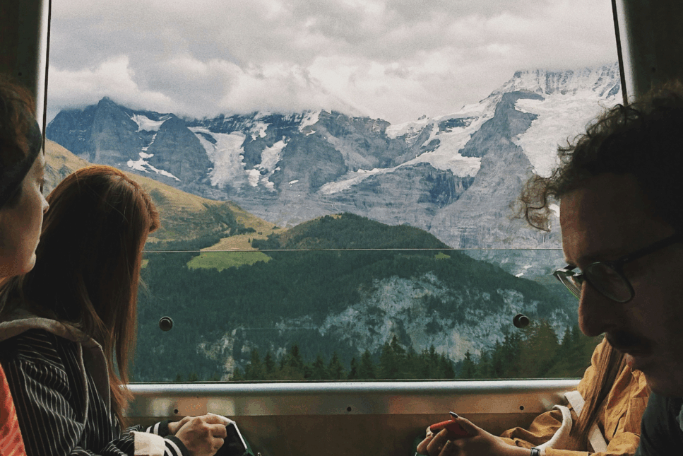 Three travelers sit inside a train with large windows, gazing at a dramatic alpine landscape of snow-capped peaks, glaciers, and green valleys outside.